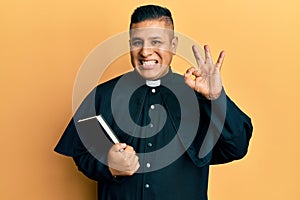 Young latin priest man holding bible doing ok sign with fingers, smiling friendly gesturing excellent symbol