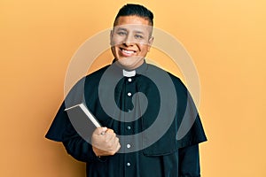 Young latin priest man holding bible looking positive and happy standing and smiling with a confident smile showing teeth