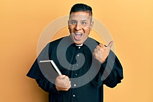 Young latin priest man holding bible pointing thumb up to the side smiling happy with open mouth