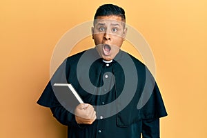 Young latin priest man holding bible scared and amazed with open mouth for surprise, disbelief face