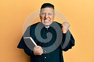 Young latin priest man holding bible screaming proud, celebrating victory and success very excited with raised arm