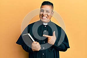 Young latin priest man holding bible smiling happy pointing with hand and finger