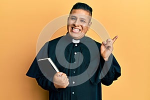 Young latin priest man holding bible smiling happy pointing with hand and finger to the side