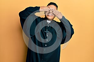 Young latin priest man standing over yellow background covering eyes with hands smiling cheerful and funny