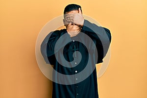 Young latin priest man standing over yellow background covering eyes and mouth with hands, surprised and shocked