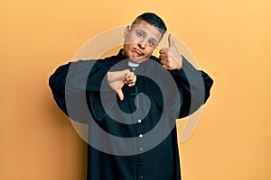 Young latin priest man standing over yellow background doing thumbs up and down, disagreement and agreement expression