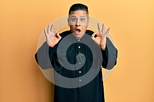 Young latin priest man standing over yellow background looking surprised and shocked doing ok approval symbol with fingers