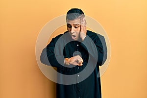 Young latin priest man standing over yellow background looking at the watch time worried, afraid of getting late