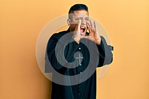 Young latin priest man standing over yellow background shouting angry out loud with hands over mouth