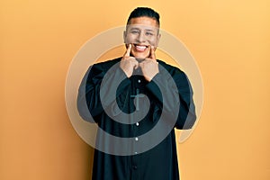 Young latin priest man standing over yellow background smiling with open mouth, fingers pointing and forcing cheerful smile