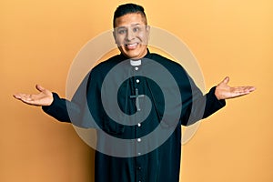 Young latin priest man standing over yellow background smiling showing both hands open palms, presenting and advertising