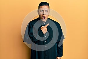 Young latin priest man standing over yellow background surprised pointing with finger to the side, open mouth amazed expression