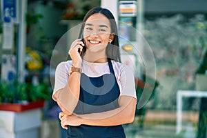 Young latin shopkeeper girl smiling happy talking on the smartphone at florist