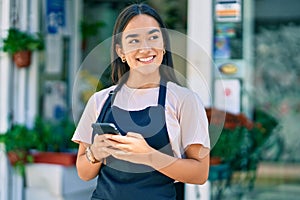 Young latin shopkeeper girl smiling happy using smartphone at florist