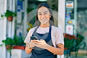 Young latin shopkeeper girl smiling happy using smartphone at florist