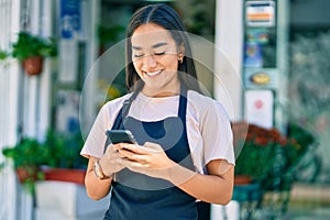 Young latin shopkeeper girl smiling happy using smartphone at florist