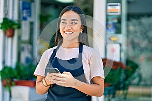 Young latin shopkeeper girl smiling happy using smartphone at florist