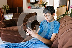 Young latin teenager with phone sitting on sofa at home in Latin America