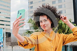 Young latin woman doing a selfie portrait with a cellphone touching her curly hair. Happy hismanic girl taking a photo