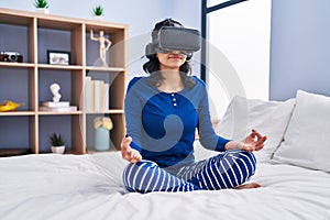 Young latin woman doing yoga exercise using virtual reality glasses at bedroom
