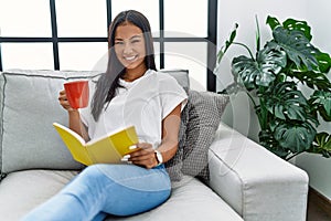 Young latin woman drinking coffee reading book at home