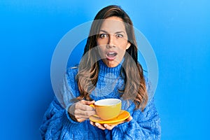 Young latin woman drinking a cup of coffee afraid and shocked with surprise and amazed expression, fear and excited face
