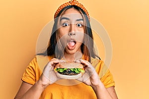 Young latin woman eating a tasty classic burger afraid and shocked with surprise and amazed expression, fear and excited face