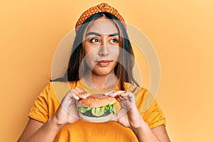 Young latin woman eating a tasty classic burger smiling looking to the side and staring away thinking