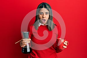 Young latin woman holding coffee filter and cup afraid and shocked with surprise and amazed expression, fear and excited face