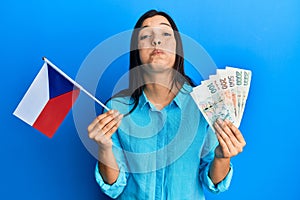 Young latin woman holding czech republic flag and koruna banknotes puffing cheeks with funny face