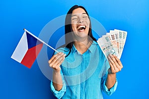 Young latin woman holding czech republic flag and koruna banknotes smiling and laughing hard out loud because funny crazy joke
