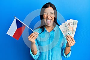 Young latin woman holding czech republic flag and koruna banknotes sticking tongue out happy with funny expression