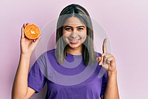 Young latin woman holding fresh orange slice smiling with an idea or question pointing finger with happy face, number one