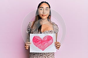 Young latin woman holding heart draw smiling looking to the side and staring away thinking