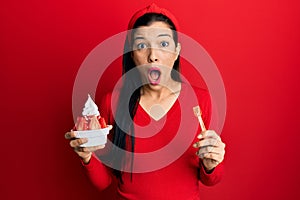 Young latin woman holding ice cream afraid and shocked with surprise and amazed expression, fear and excited face
