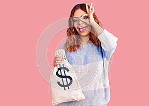 Young latin woman holding money bag with dollar symbol smiling happy doing ok sign with hand on eye looking through fingers