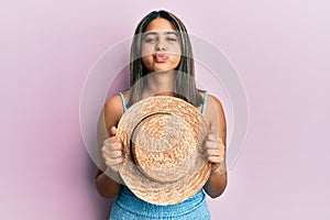 Young latin woman holding summer hat puffing cheeks with funny face