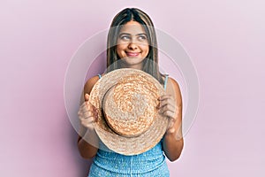 Young latin woman holding summer hat smiling looking to the side and staring away thinking