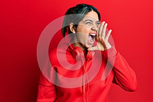 Young latin woman listening to music using headphones shouting and screaming loud to side with hand on mouth