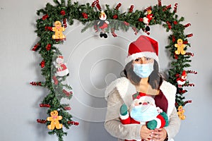 Young latin woman with protection mask, sweater and santa claus hat, christmas decoration and santa claus doll, new normal covid-1