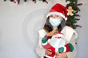 Young latin woman with protection mask, sweater and santa claus hat, christmas decoration and santa claus doll, new normal covid-1