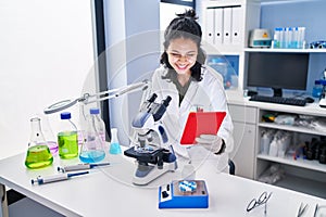 Young latin woman scientist using microscope and touchpad at laboratory