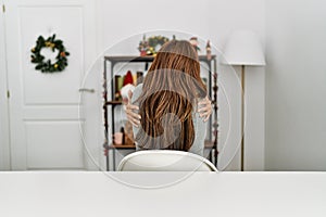 Young latin woman sitting on the table by christmas decor hugging oneself happy and positive from backwards