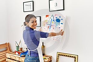 Young latin woman smiling confident hanging draw canvas on wall at art studio