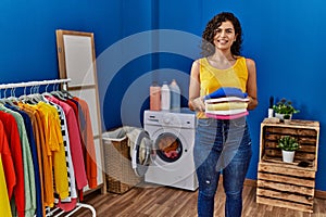 Young latin woman smiling confident holding folded clothes at laundry room