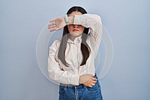 Young latin woman standing over blue background covering eyes with arm, looking serious and sad