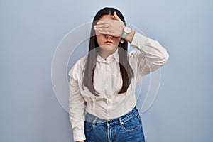 Young latin woman standing over blue background covering eyes with hand, looking serious and sad