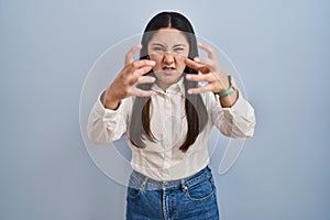 Young latin woman standing over blue background shouting frustrated with rage, hands trying to strangle, yelling mad