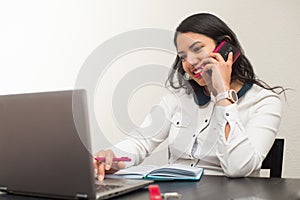 Young latin woman talking by mobile phone and working at home with the laptop. Teleworking concept on a white background