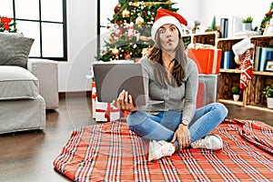 Young latin woman using laptop sitting by christmas tree making fish face with lips, crazy and comical gesture